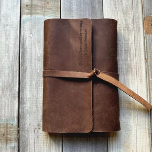 May include: A closed, brown leather journal with a strap closure. The journal has the text "Do Not Open until 07.17.2034 B. Jason Swanson" embossed on the front cover. The journal is lying on a weathered wooden surface.