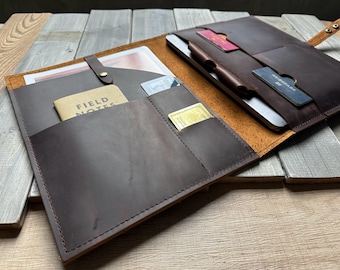 May include: A dark brown leather portfolio with multiple pockets and compartments. It features a small notebook labeled "FIELD NOTES", slots for cards, and a pen holder. The portfolio is open, revealing a tablet and other items.