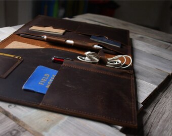 May include: A dark brown leather folio with multiple pockets and compartments. It contains a small notepad, pens, a credit card, and a blue notebook labeled "FIELD NOTES". A white charging cable is secured with a leather strap.