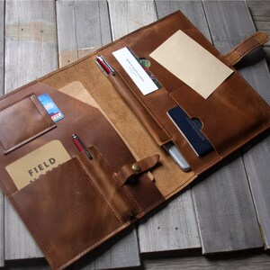 May include: Open brown leather portfolio with multiple pockets, holding pens, cards, and a notepad. The portfolio is open, revealing a light brown interior and a snap closure. The background is a rustic wooden surface.