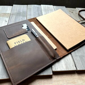 May include: Open brown leather journal with a card slot, pen holder, and a small notebook labeled "FIELD NOTES." The journal is open, revealing a blank notepad. A silver pen is in the holder. The journal is on a wooden surface.