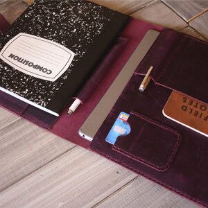 May include: A deep purple leather portfolio with a composition notebook, a tablet, and a small notebook labeled "FIELD NOTES." The portfolio has pockets for cards and pens. The notebook has a white label with the word "COMPOSITION."