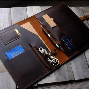 Könnte beinhalten: Geöffnete dunkelbraune Ledermappe mit mehreren Fächern für Karten, Stifte und einen Notizblock. Das Innere ist hellbraun. Ein Stift und eine blaue Karte mit der Aufschrift "FIELD NOTES" sind sichtbar. Auf einer Holzoberfläche platziert.