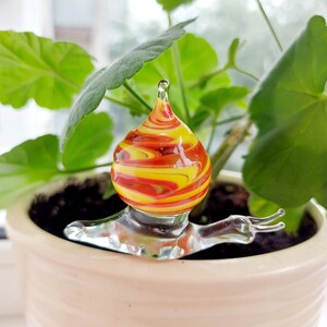 Puede incluir: Un colorido globo de riego para plantas con forma de caracol de cristal. El globo tiene forma de lágrima con patrones arremolinados de amarillo, naranja y rojo. El cuerpo del caracol es de cristal transparente, en una maceta de cerámica beige.