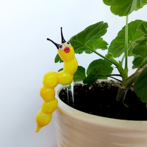 May include: A whimsical, yellow glass caterpillar plant watering stake with black antennae and red lips. The decorative garden art is placed in a beige pot with green leaves. The caterpillar is approximately 13 cm long.