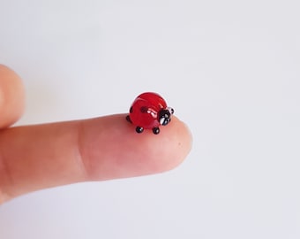 Può includere: Una minuscola statuina a forma di coccinella, prevalentemente rossa con pois neri e testa nera. La coccinella è appoggiata su un dito, evidenziando le sue piccole dimensioni e i dettagli delicati. La statuina è probabilmente realizzata in vetro o materiale simile.