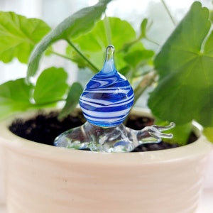 May include: A glass plant watering globe with a blue and white striped bulb and clear glass base. The globe is placed in a beige ceramic pot with green leafy plants in the background. The watering globe is designed to slowly release water to the plant.