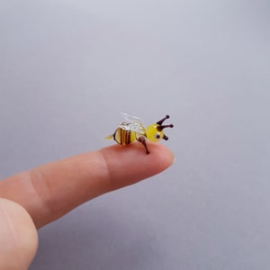 May include: A small glass bee figurine with yellow and brown stripes, clear wings, and black eyes. The bee is perched on a finger.