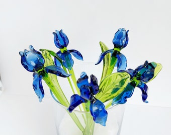 May include: A clear glass vase holds a collection of blue glass iris flowers with green stems and leaves. The flowers are a vibrant blue, and the leaves have a touch of yellow-green. The vase sits on a white surface.