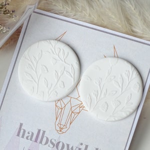 Puede incluir: Un par de pendientes de cerámica blanca con un diseño floral. Los pendientes son redondos y tienen un diseño en relieve de hojas y flores. Los pendientes están en una tarjeta blanca con el texto "halbsowild jewellery".