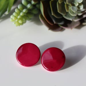 May include: A pair of round, glossy, deep red earrings. The earrings are displayed on a white surface, with a succulent and other greenery in the background. The earrings are a simple, classic design.