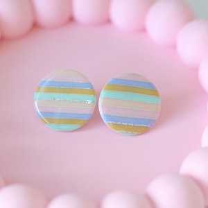 Pode incluir: Dois brincos redondos com um design de listras pastel em tons de rosa, azul, dourado e verde menta. Os brincos são exibidos em uma superfície rosa, cercados por uma borda de contas rosa.