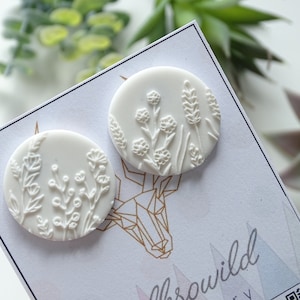 Puede incluir: Dos pendientes de cerámica blanca con un diseño floral en relieve. Los pendientes son redondos y tienen un fondo blanco con un patrón floral blanco.