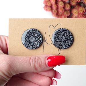 Puede incluir: Pendientes de botón redondos negros con un diseño floral plateado. Se muestran en una tarjeta marrón con una firma manuscrita. El fondo incluye flores rosas. Diámetro aproximado: 2.5 cm.