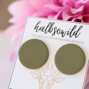 Puede incluir: Pendientes de botón redondos de color verde oliva presentados en una tarjeta blanca con el texto "halbso wild" y "THE BEAUTY OF HANDMADE JEWELRY". Los pendientes se colocan sobre un fondo floral rosa borroso.