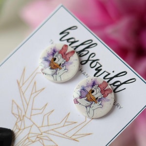 May include: A pair of round Daisy Duck earrings. Each earring features a watercolor-style image of Daisy Duck with a pink bow and purple accents. The earrings are on a white card with gold geometric designs and the text "halbsowild".