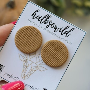 May include: Two circular tan stud earrings with a textured, grid-like pattern on a white display card. The card features the text "halbswild" and "THE BEAUTY OF HANDMADE JEWELRY" along with a geometric deer illustration and a QR code.