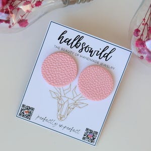 Könnte beinhalten: Ein Paar hellrosa, strukturierte, runde Ohrringe, die auf einer weißen Karte präsentiert werden. Auf der Karte steht "halbsowild" und "THE BEAUTY OF HANDMADE JEWELRY". Darunter ein geometrisches Hirschkopf-Design und die Worte "perfectly imperfect".