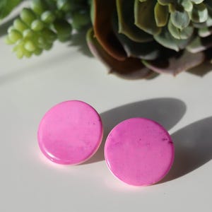 May include: Two bright pink, round earrings with a glossy finish are displayed on a white surface. The earrings are the focal point, with a succulent plant in the background. The earrings cast a shadow.