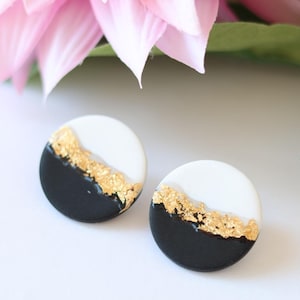 Könnte beinhalten: Ein Paar runde Ohrstecker. Jeder Ohrring ist halb schwarz und halb weiß, mit Akzenten aus Blattgold. Die Ohrringe sind vor einem weißen Hintergrund platziert, mit rosa Blüten und grünen Blättern im Hintergrund.