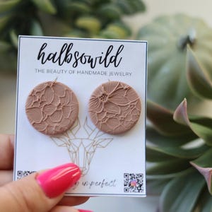 May include: Two circular, dusty rose-coloured stud earrings featuring a raised, intricate floral lace texture. The earrings are mounted on a white display card printed with the text "halbsowild THE BEAUTY OF HANDMADE JEWELRY" and a geometric deer head graphic.