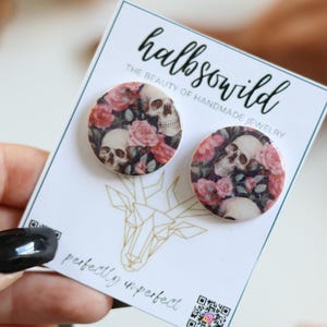 Puede incluir: Pendientes redondos con un diseño de calavera y rosas. Los pendientes se muestran en una tarjeta blanca con el texto "halbso wild" y "perfectly imperfect". Los pendientes tienen un fondo oscuro con rosas rosas y calaveras blancas.