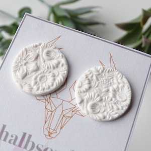 Könnte beinhalten: Ein Paar weiße Keramikohrringe mit einem floralen Design. Die Ohrringe sind rund und haben ein erhabenes Design.