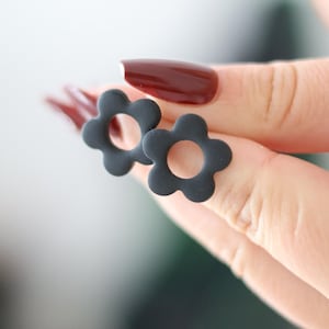 Peut inclure: Une paire de boucles d'oreilles à clous noir mat en forme de fleurs à cinq pétales avec une découpe circulaire au centre de chacune. Les boucles d'oreilles présentent une finition lisse et unie, soulignant la silhouette florale distincte.