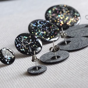 May include: A set of five black stud earrings with silver glitter embedded in the resin. The earrings are different sizes and are arranged in a row from largest to smallest.