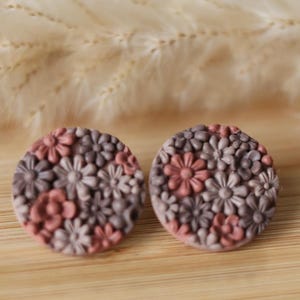 Puede incluir: Un par de pendientes de botón redondos con un diseño floral. Los pendientes presentan un patrón de pequeñas flores en tonos malva y rosa empolvado. Se encuentran sobre una superficie de madera.