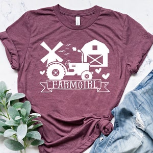 May include: A maroon t-shirt with a white graphic of a tractor, windmill, and barn, with the text "FARMGIRL" on a banner. The shirt is paired with blue jeans. A sprig of green leaves is in the lower left corner.