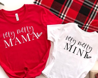 Op de afbeelding: Twee rood-witte kersttruien met de tekst "very merry mama" en "very merry mini" erop. De truien liggen op een wit oppervlak met een rood-zwart geruite deken op de achtergrond.