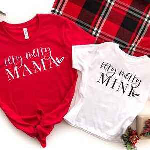 May include: Two red and white Christmas shirts with the text "very merry mama" and "very merry mini" on them. The shirts are laid out on a white surface with a red and black plaid blanket in the background.