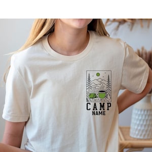 May include: A cream coloured t-shirt with a graphic of a camping scene with a coffee mug and a tent. The text "CAMP NAME" is below the graphic.