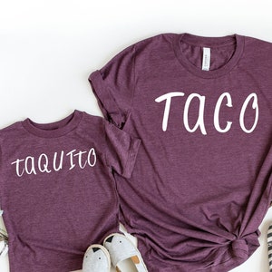 May include: Two maroon t-shirts with the words "Taco" and "Taquito" printed in white. The shirts are laid out on a white background with some greenery and cotton.