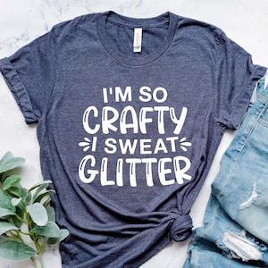 Może przedstawiać: Koszulka w ciemnoniebieskim kolorze z napisem "I'm so Crafty I Sweat Glitter" w kolorze białym.
