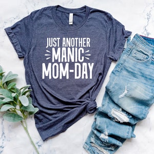 May include: A dark blue t-shirt with the text "Just Another Manic Mom-Day" printed in white on the front. The shirt is laid out on a white marble surface with a pair of blue jeans folded next to it.