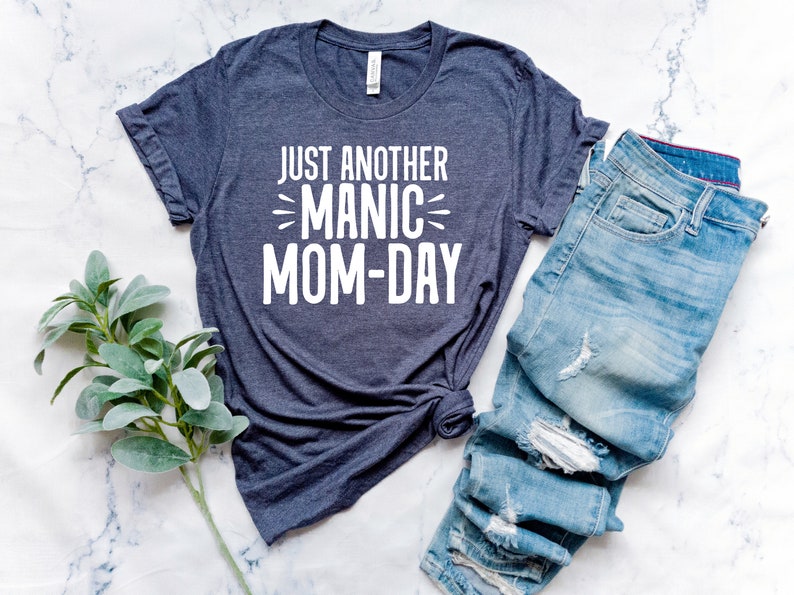 May include: A dark blue t-shirt with the text "Just Another Manic Mom-Day" printed in white on the front. The shirt is laid out on a white marble surface with a pair of blue jeans folded next to it.