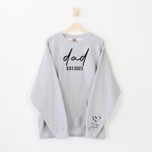 K&ouml;nnte beinhalten: Grauer Pullover mit dem Text "dad est. 2023" auf der Vorderseite und einem Herz mit dem Text "mary jane" auf dem &Auml;rmel.