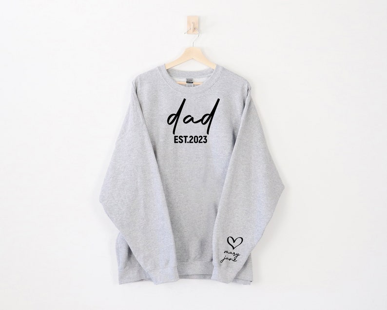 K&ouml;nnte beinhalten: Grauer Pullover mit dem Text "dad est. 2023" auf der Vorderseite und einem Herz mit dem Text "mary jane" auf dem &Auml;rmel.