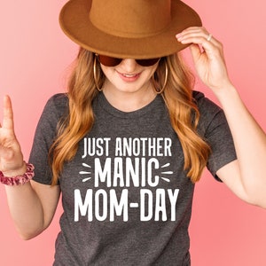 May include: A woman wearing a gray t-shirt with the text "Just Another Manic Mom-Day" in white lettering. She is wearing a brown hat and sunglasses and is holding her hat with one hand and giving a peace sign with the other.