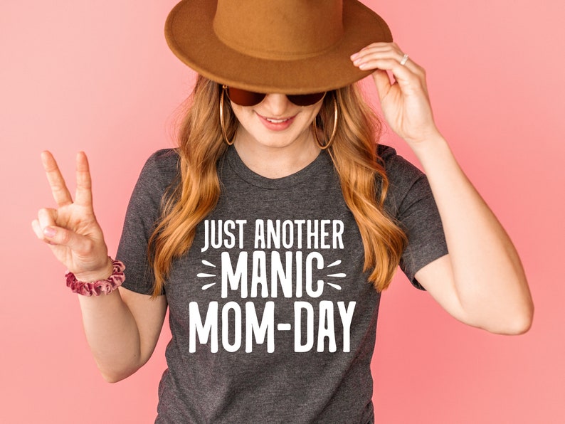 May include: A woman wearing a gray t-shirt with the text "Just Another Manic Mom-Day" in white lettering. She is wearing a brown hat and sunglasses and is holding her hat with one hand and giving a peace sign with the other.