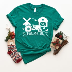 May include: Teal t-shirt with a white graphic featuring a tractor, windmill, barn, and the word "FARMGIRL" on a banner. The shirt is styled with a knot at the bottom and is surrounded by holiday decorations, including a patterned sock and wrapped gifts.