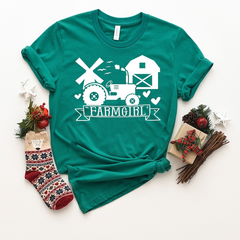 May include: Teal t-shirt with a white graphic featuring a tractor, windmill, barn, and the word "FARMGIRL" on a banner. The shirt is styled with a knot at the bottom and is surrounded by holiday decorations, including a patterned sock and wrapped gifts.