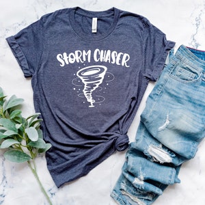 May include: A dark blue t-shirt with a white graphic of a tornado and the text "Storm Chaser". The shirt is laid out on a white marble surface with a pair of blue jeans.