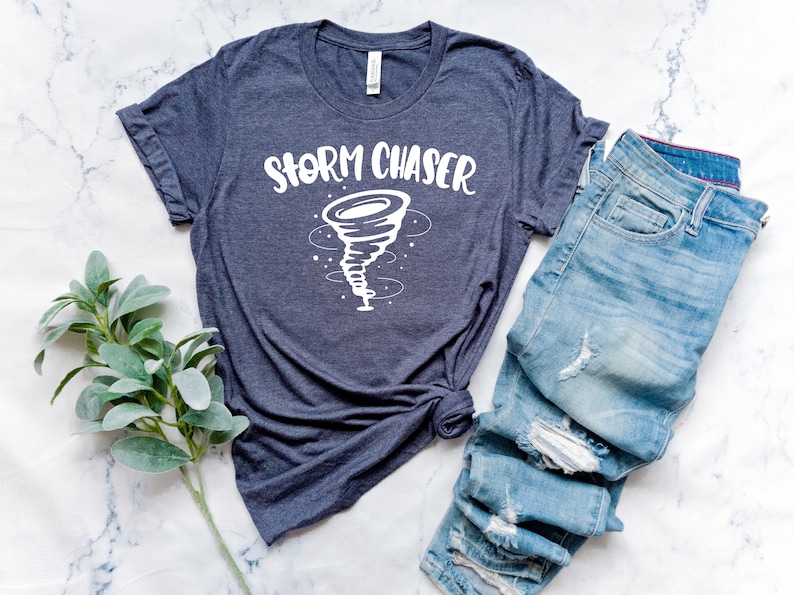 May include: A dark blue t-shirt with a white graphic of a tornado and the text "Storm Chaser". The shirt is laid out on a white marble surface with a pair of blue jeans.