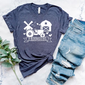 May include: A navy blue t-shirt with a white graphic of a tractor, windmill, barn, and the text "FARMGIRL". The shirt is paired with blue denim jeans. The shirt is made of a soft material.