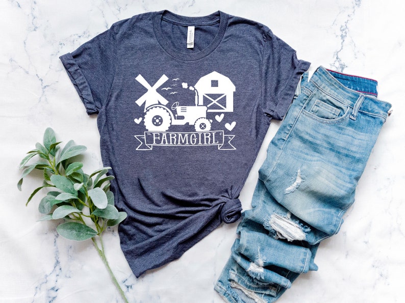 May include: A navy blue t-shirt with a white graphic of a tractor, windmill, barn, and the text "FARMGIRL". The shirt is paired with blue denim jeans. The shirt is made of a soft material.