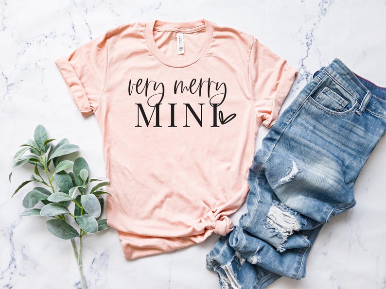 May include: A peach-colored t-shirt with the text "very merry mini" in black lettering with a small heart.