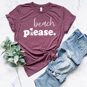 May include: A maroon t-shirt with the words "beach please" in white script and block letters. The shirt is paired with distressed blue jeans and a sprig of green leaves, all arranged on a white marble surface.